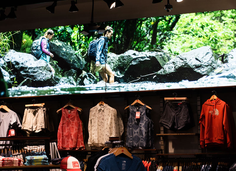 inside of a store, above some clothes on racks is a photo of a man and woman hiking through the woods, over some rocks in a river